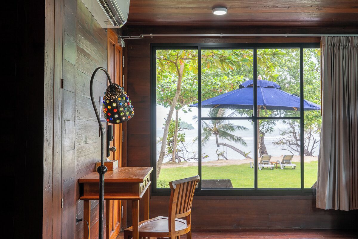 A wooden interior with a table and chair, a window with a view of a beach and umbrellas, and a curtain on the window.