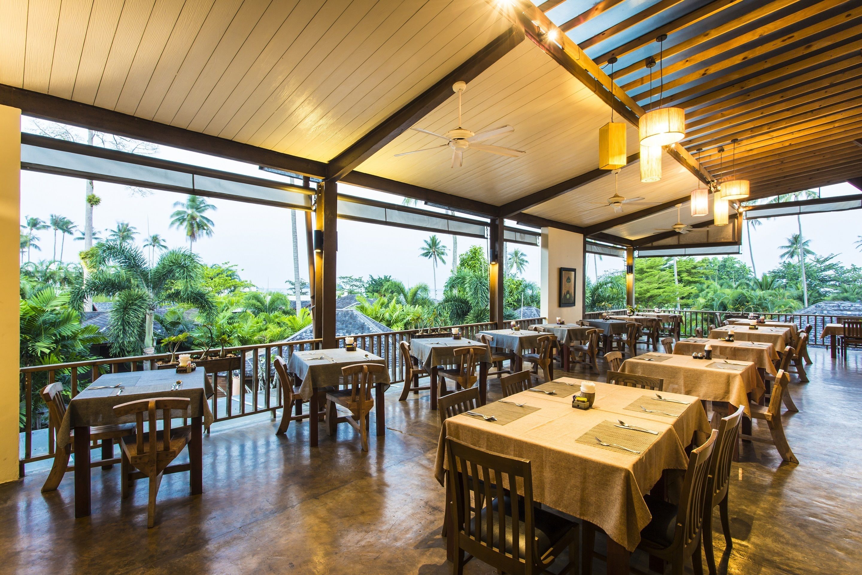 Un restaurante amplio con mesas y sillas de madera, ventanas grandes y vistas a palmeras.