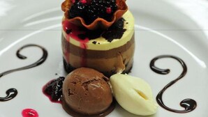 Un dessert composé d’un gâteau à plusieurs couches, de glace au chocolat et d’une boule de glace à la vanille, le tout présenté sur une assiette blanche et décoré de filaments de chocolat.