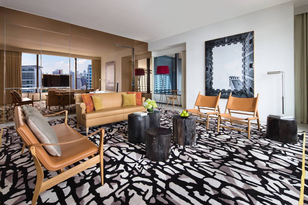 A modern hotel room with a large window, a sofa, two armchairs, a coffee table, and a patterned rug.