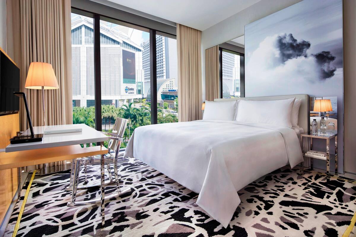 A hotel room with a large bed, a desk with a lamp, a chair, and a view of the cityscape through large windows.