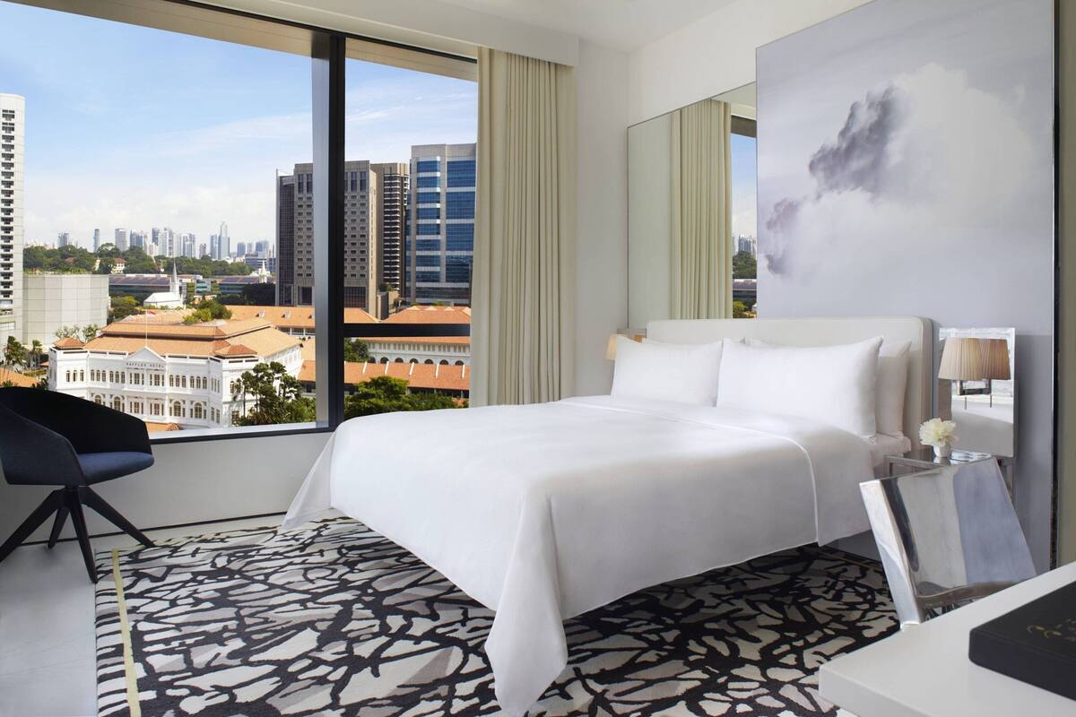 A hotel room with a large bed, a view of the cityscape, a modern chair, and a decorative wall piece.