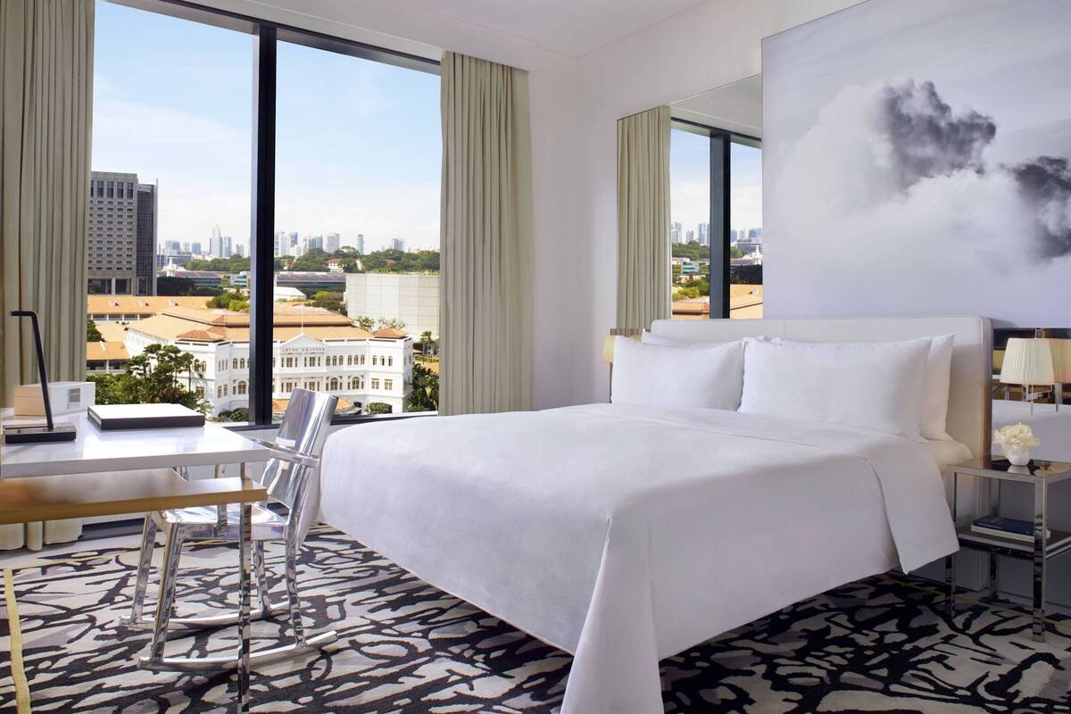 A hotel room with a large bed, a desk, a chair, and a view of the cityscape through large windows.