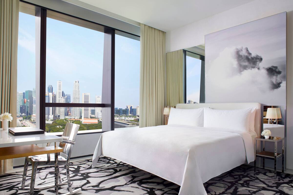A hotel room with a large bed, a desk with a chair, a view of the city skyline through large windows, and a modern abstract painting on the wall.