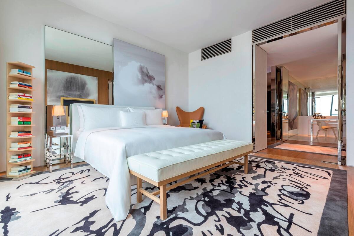 A modern hotel room with a large bed, a bedside table with a lamp, a bookshelf, and a large abstract painting on the wall.