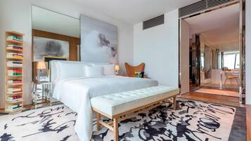 A modern hotel room with a large bed, a bedside table with a lamp, a bookshelf, and a large abstract painting on the wall.