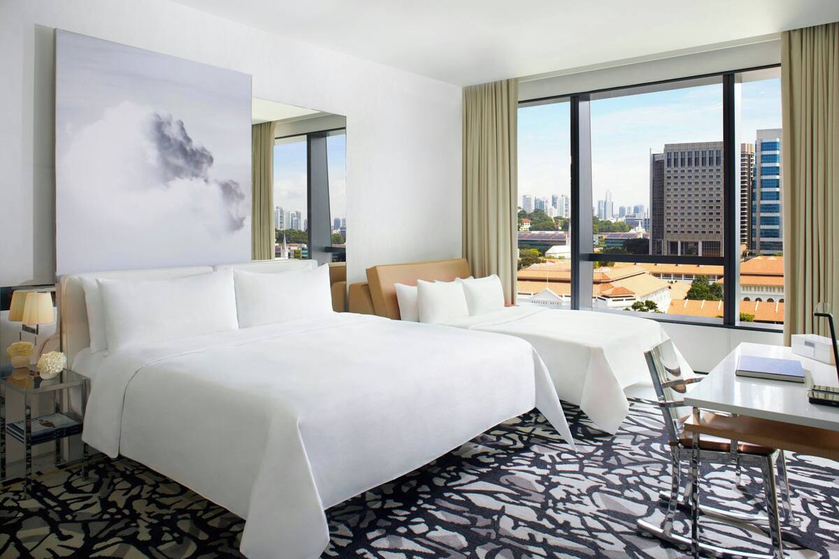 A hotel room with a large bed, a smaller bed, a desk with a chair, a nightstand with a lamp, and a view of the city through large windows.