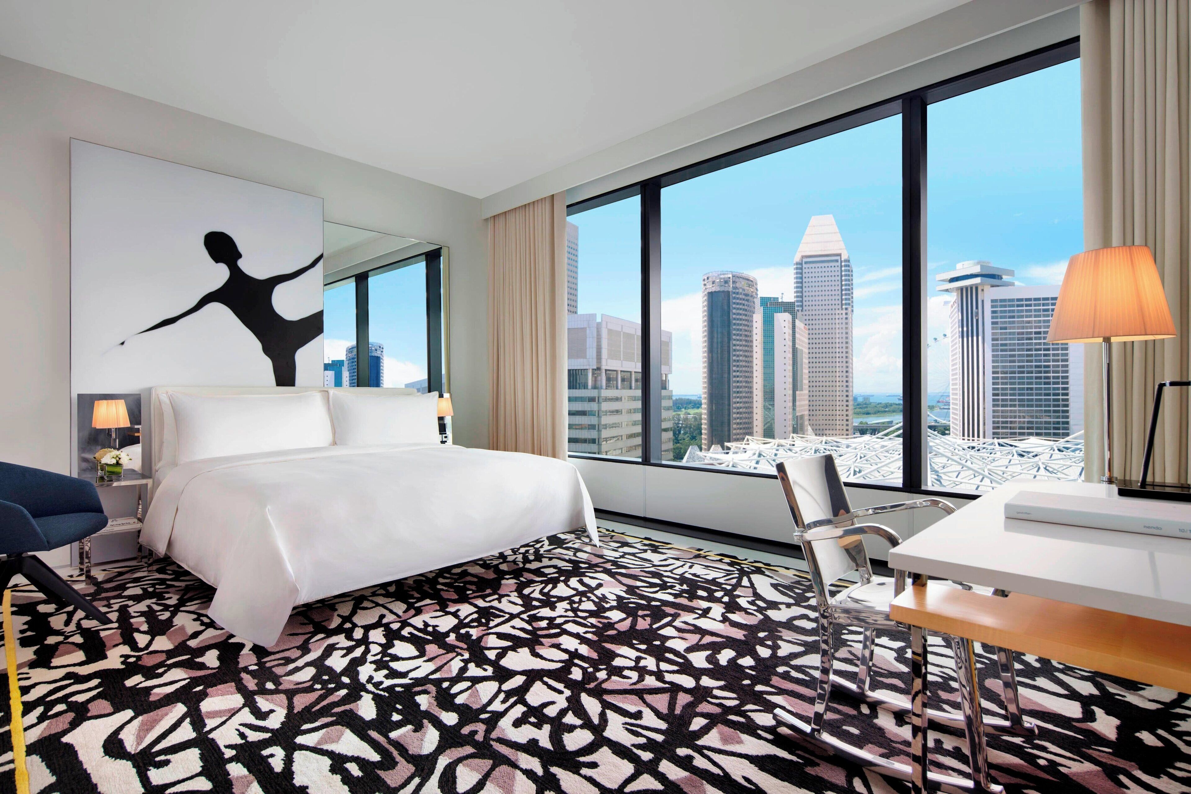 A hotel room with a large bed, a desk, a chair, and a view of the cityscape through large windows.