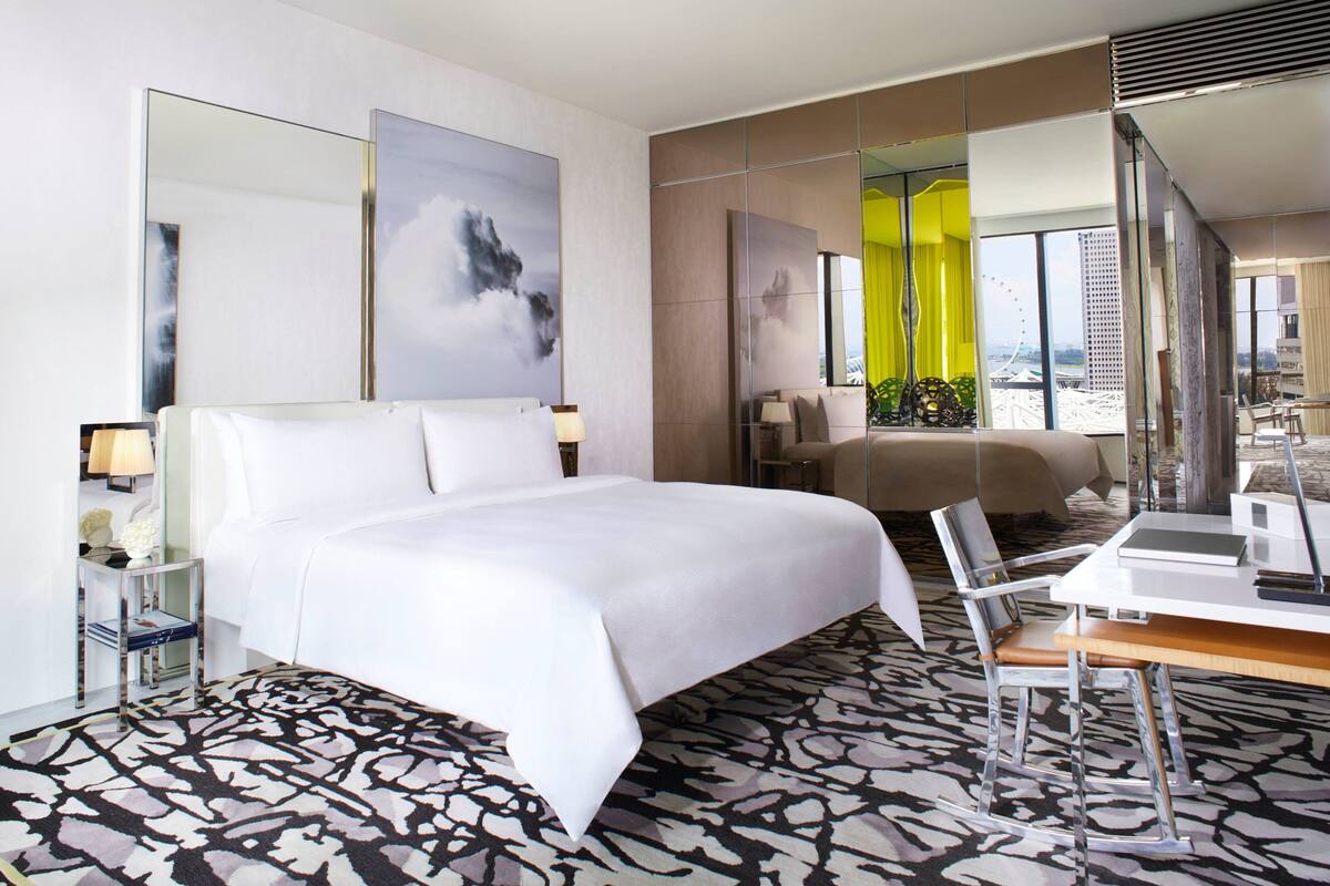 A modern hotel room with a large bed, a desk with a chair, a large mirror, and a view of a cityscape.
