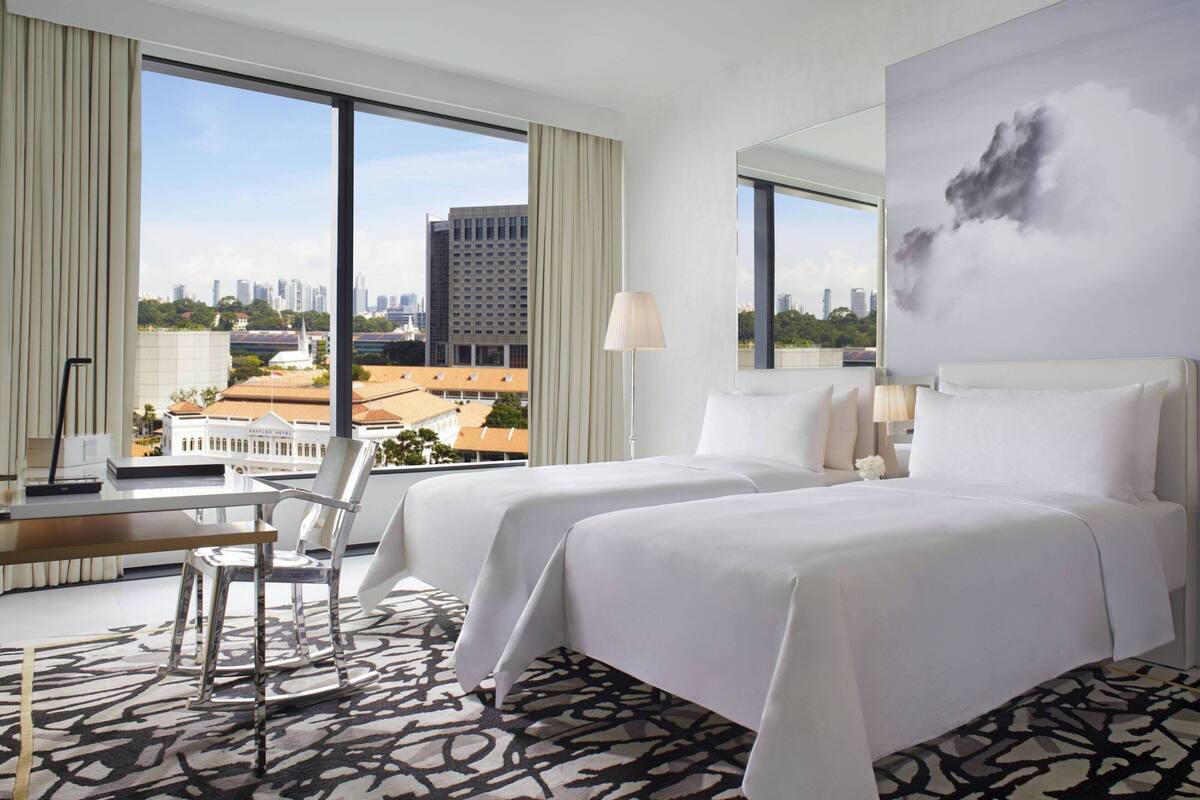 A hotel room with two beds, a desk, and a chair. There is a large window offering a city view.