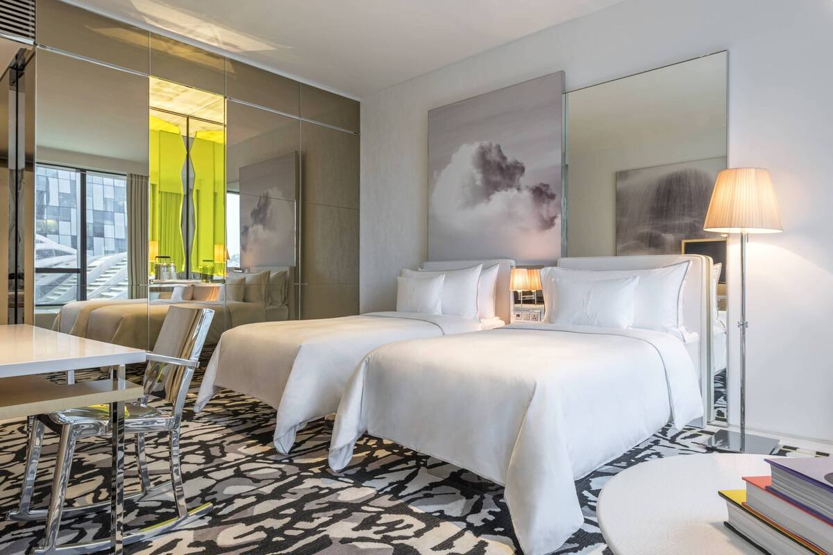 A hotel room with two beds, a dining table, chairs, a lamp, and a large window with a view of a building.