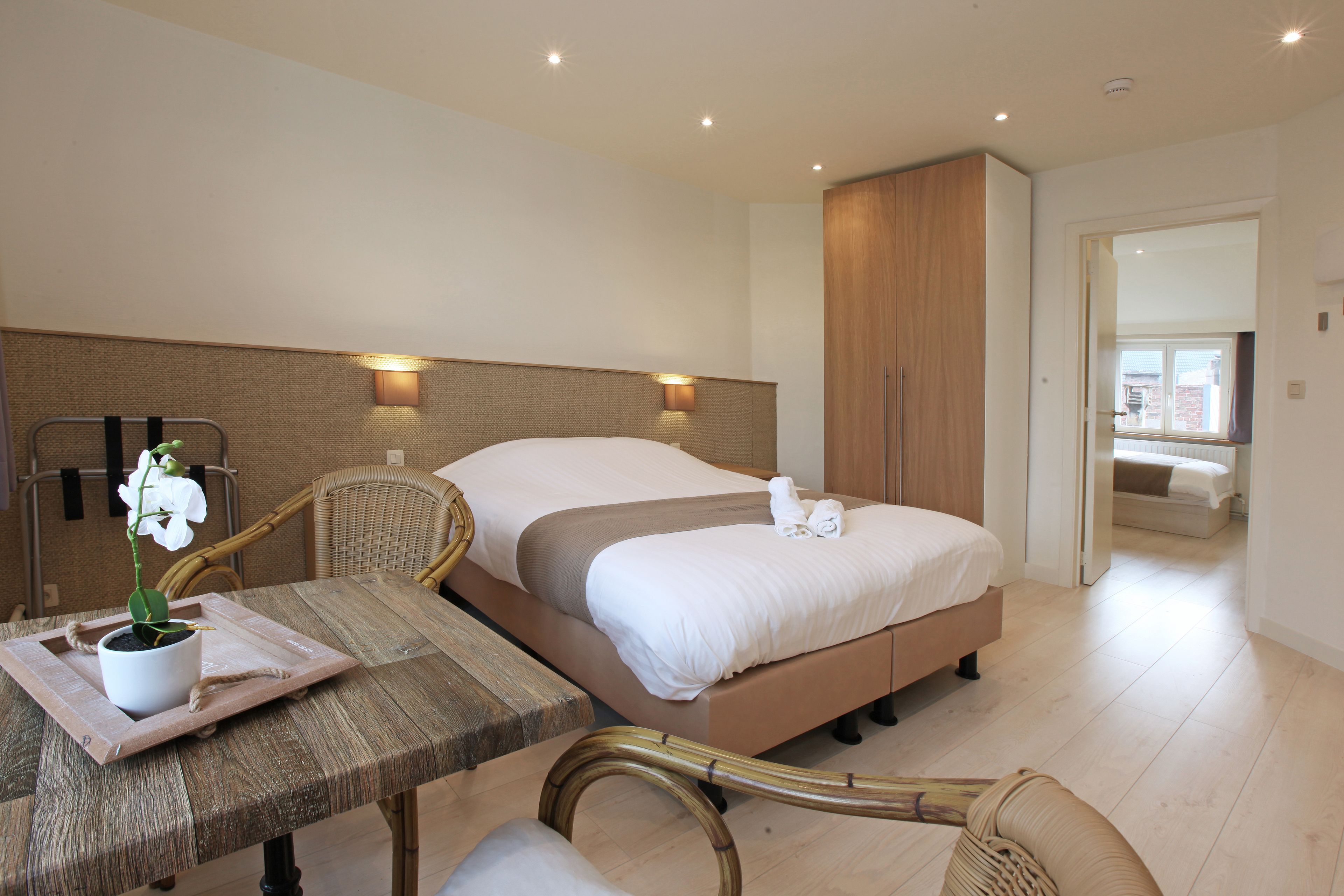 Una habitación de hotel con una cama, mesitas de noche, una planta en maceta y vista de la habitación contigua.