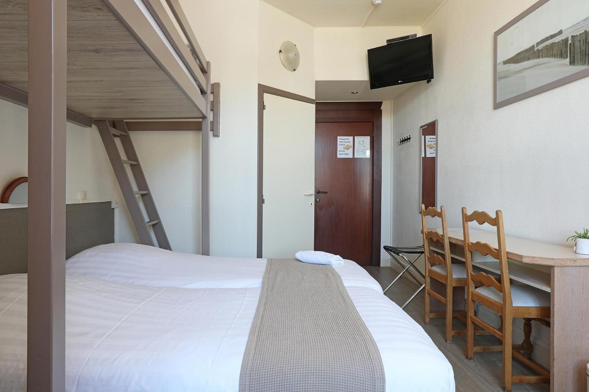 A hotel room with a bunk bed, a desk with chairs, a television, and a door with a sign.