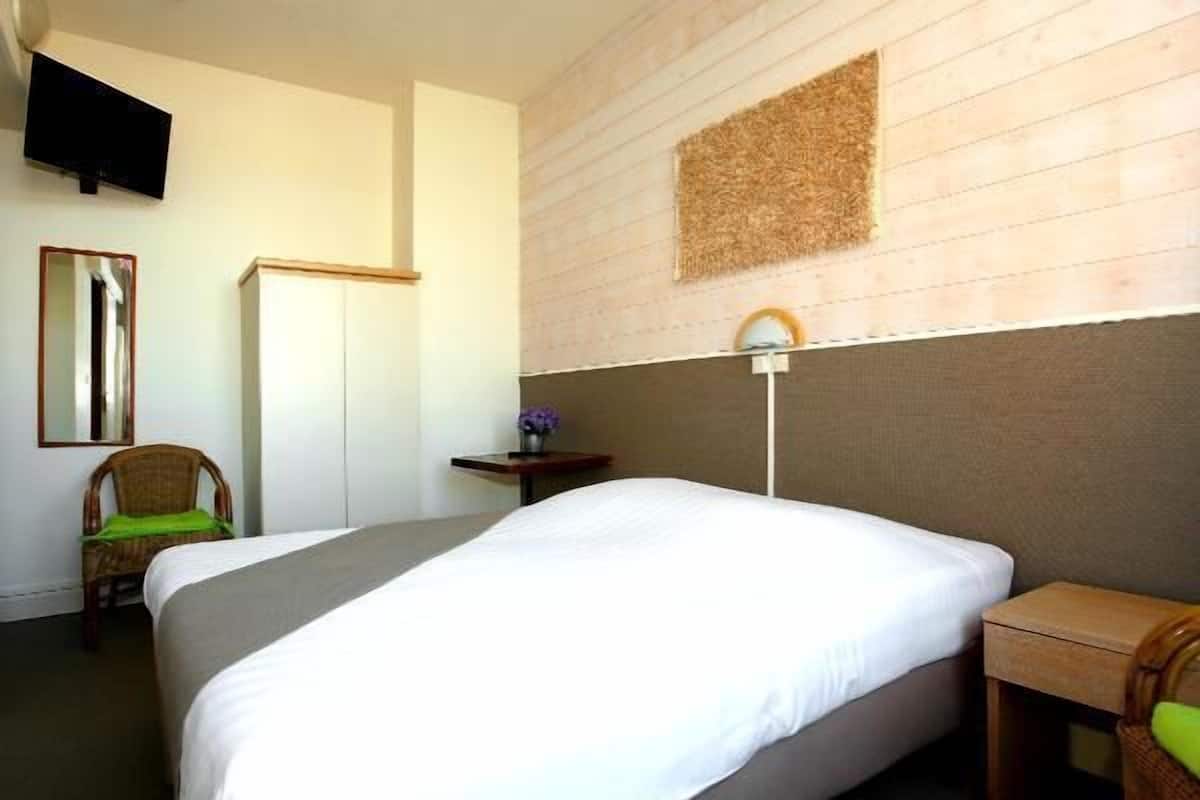 Une chambre d’hôtel avec un lit, une chaise, une petite table sur laquelle se trouve un vase, et un téléviseur fixé au mur.