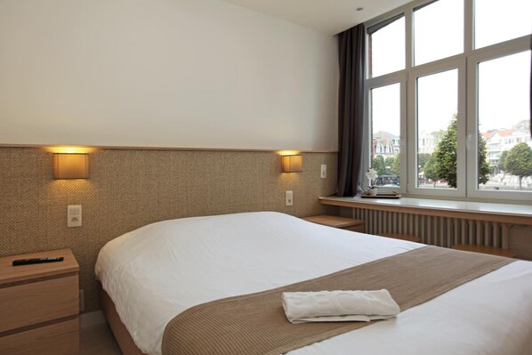 A hotel room with a large bed, two bedside lamps, a window with a view of buildings, and a radiator under the window.