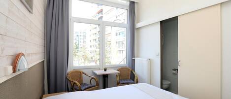 A hotel room with a bed, two wicker chairs, a small table, a window with curtains, and a view of buildings outside.