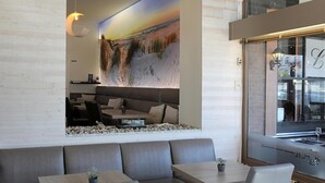 A modern interior with a large wall mural, a seating area with tables, and a view of the street through a glass window. - Hotel Cecil (De Panne)