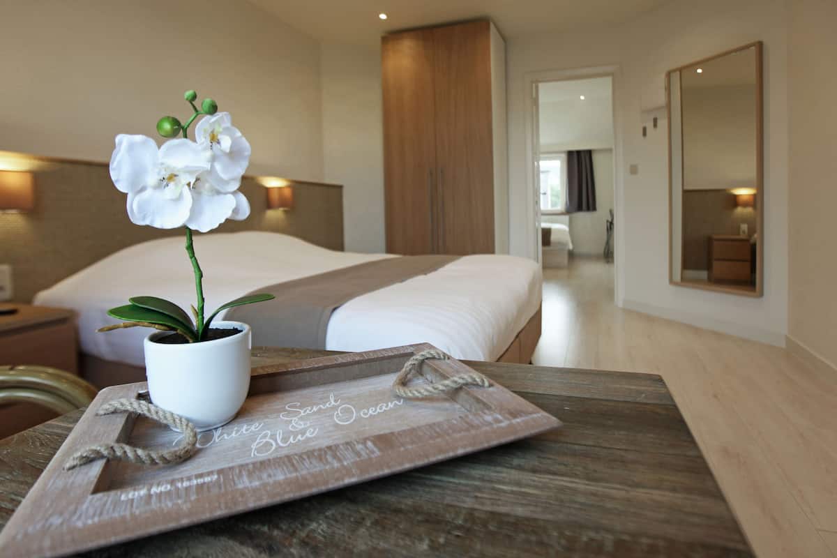 Une chambre d’hôtel avec un lit, une table de chevet en bois sur laquelle se trouve un plateau, une orchidée en pot et un miroir.