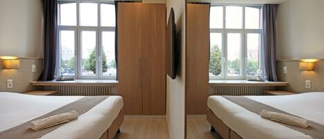A mirrored hotel room with two beds, a large window with curtains, a flat-screen TV, and a radiator under the window.