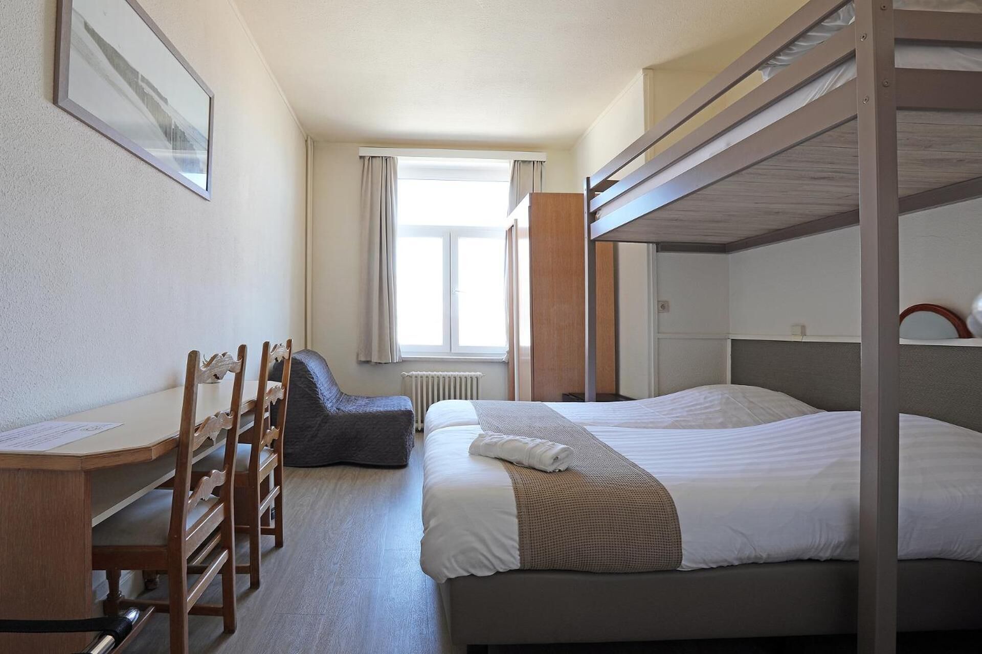 Una habitación de hotel con literas, un escritorio con sillas, una ventana con cortinas y un radiador.