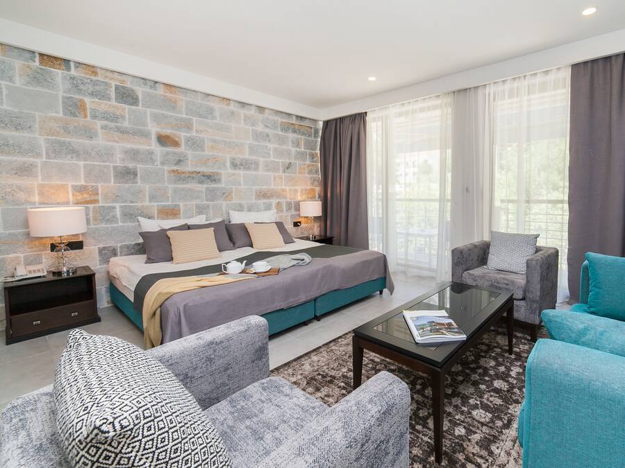 Une chambre d’hôtel moderne avec un mur d’accent en pierre, un grand lit, des meubles rembourrés aux tons de gris et de bleu, et une table basse en verre.