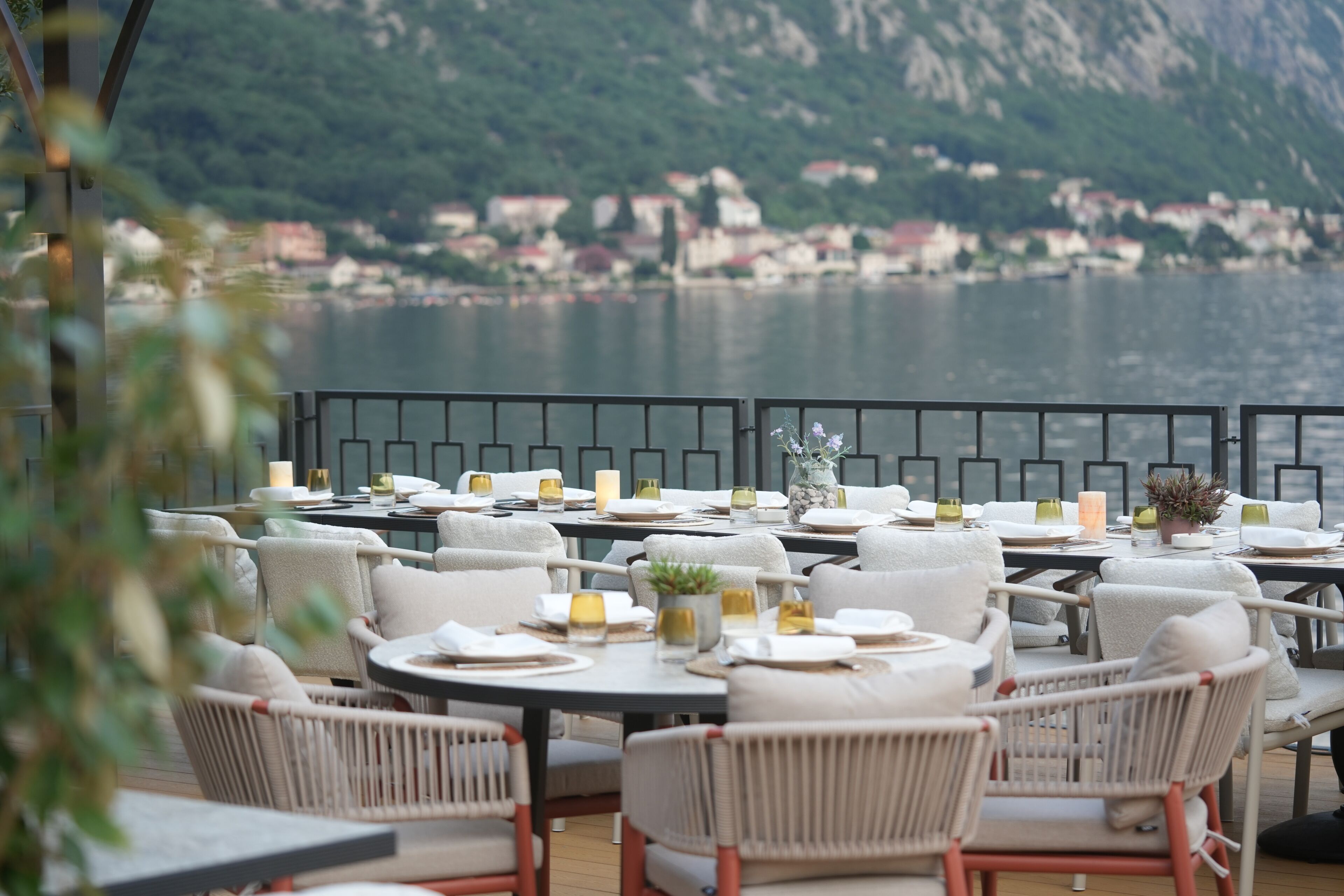 En restaurantterrasse med runde borde og stole, med udsigt over vand og et bjerglandskab.