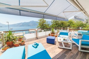 Un espacio al aire libre iluminado por el sol, con cojines azules, plantas en macetas y vistas al mar y a las montañas.