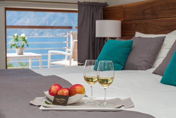 Une chambre d’hôtel avec un lit, deux verres à vin, un saladier de fruits et une vue sur la mer.