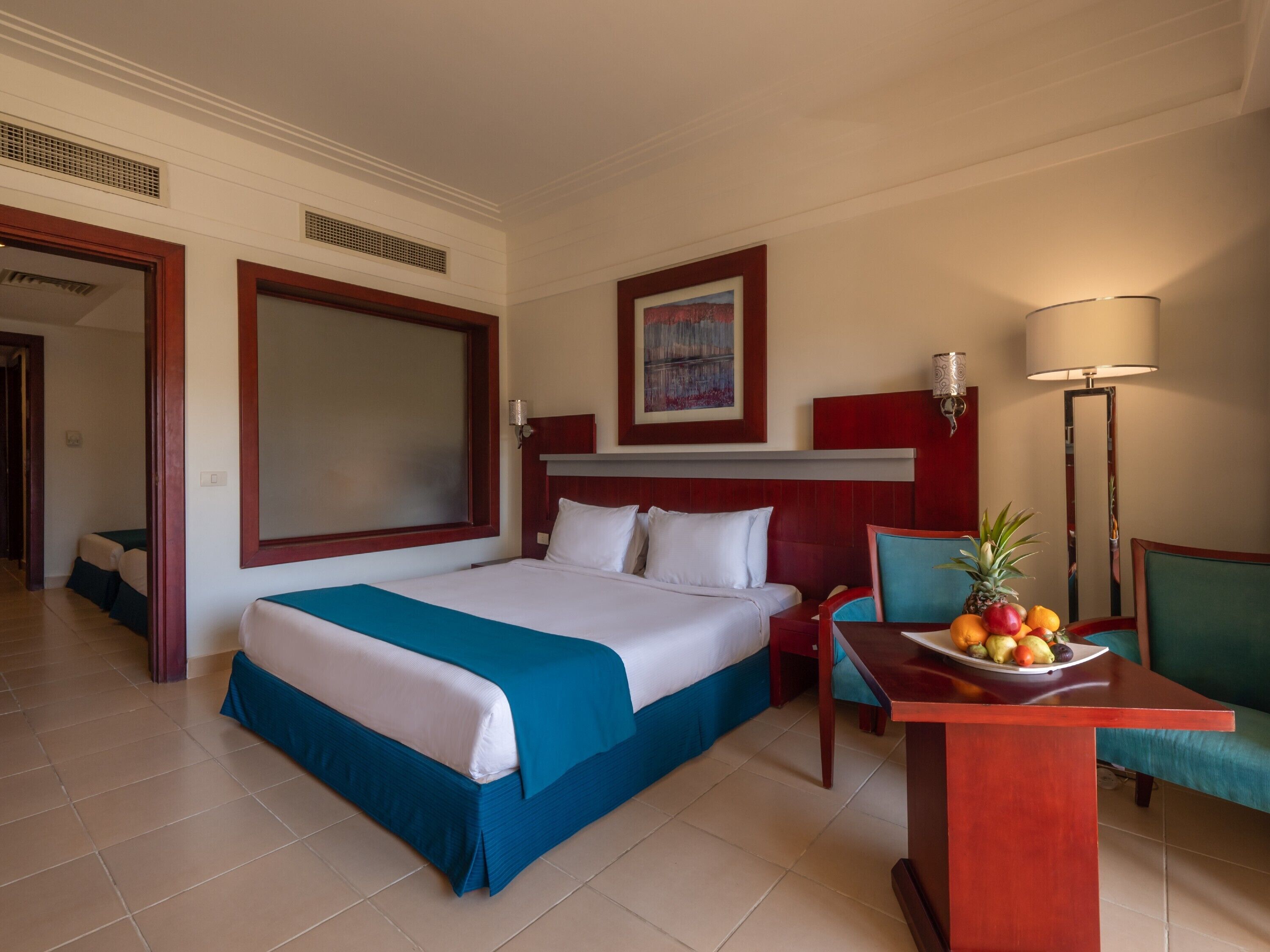 A hotel room with a large bed, a desk with a fruit plate, a chair, and a lamp.