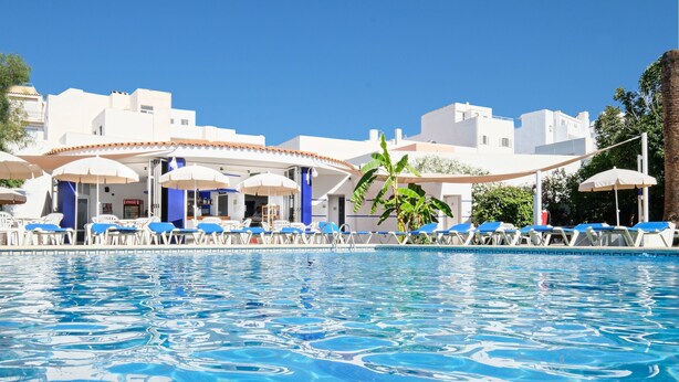 A swimming pool with lounge chairs and umbrellas, surrounded by white buildings with blue accents. - azuLine Hotel Llevant (Sant Antoni de Portmany)
