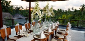 A table set for a meal with white flowers and clear glassware, overlooking a tropical landscape with palm trees.