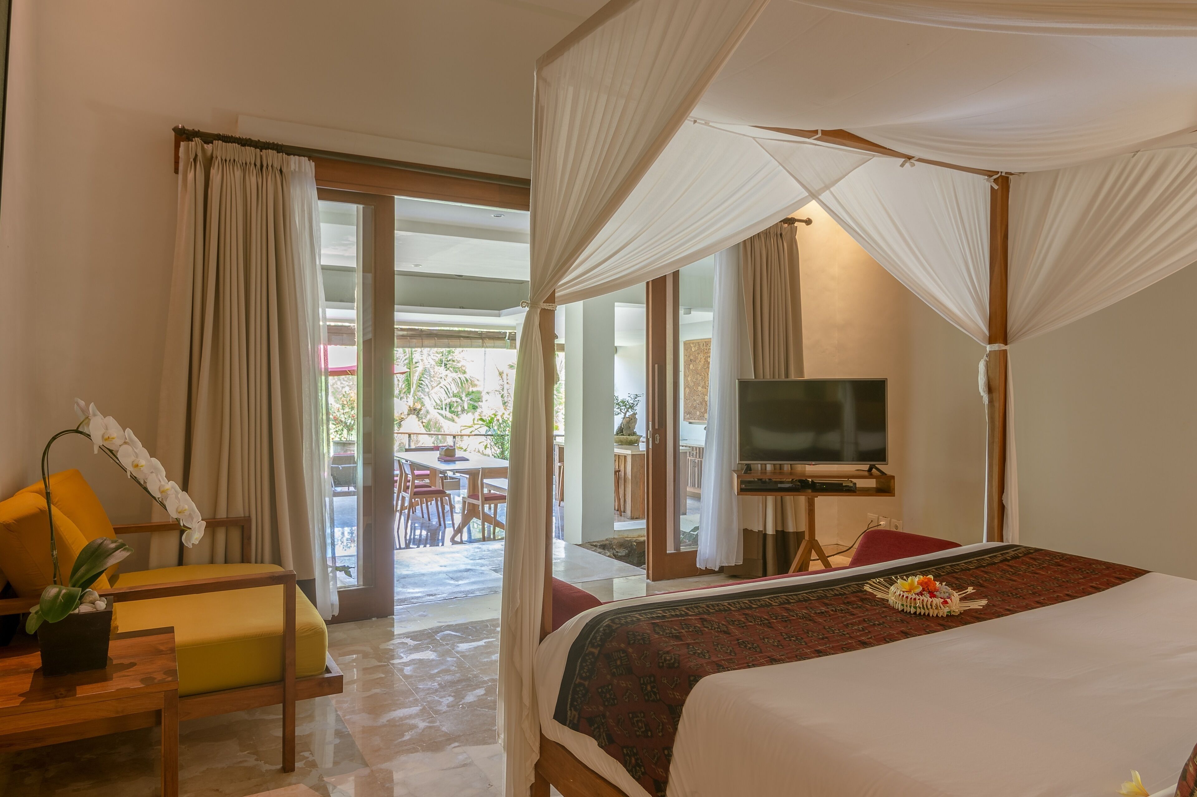 Quarto de hotel com cama, televisão, área de refeições com cadeiras e mesa, e varanda com cortinas.