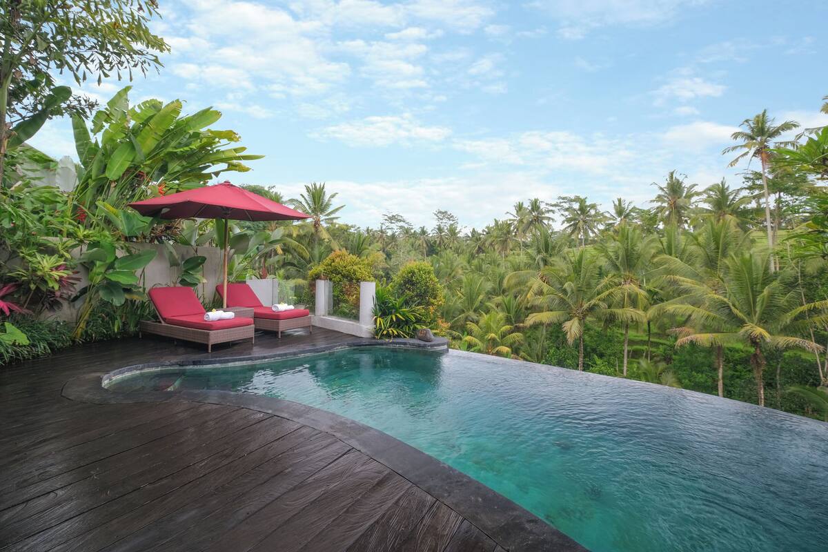 A infinity pool with red lounge chairs and a red umbrella, surrounded by lush greenery and palm trees.