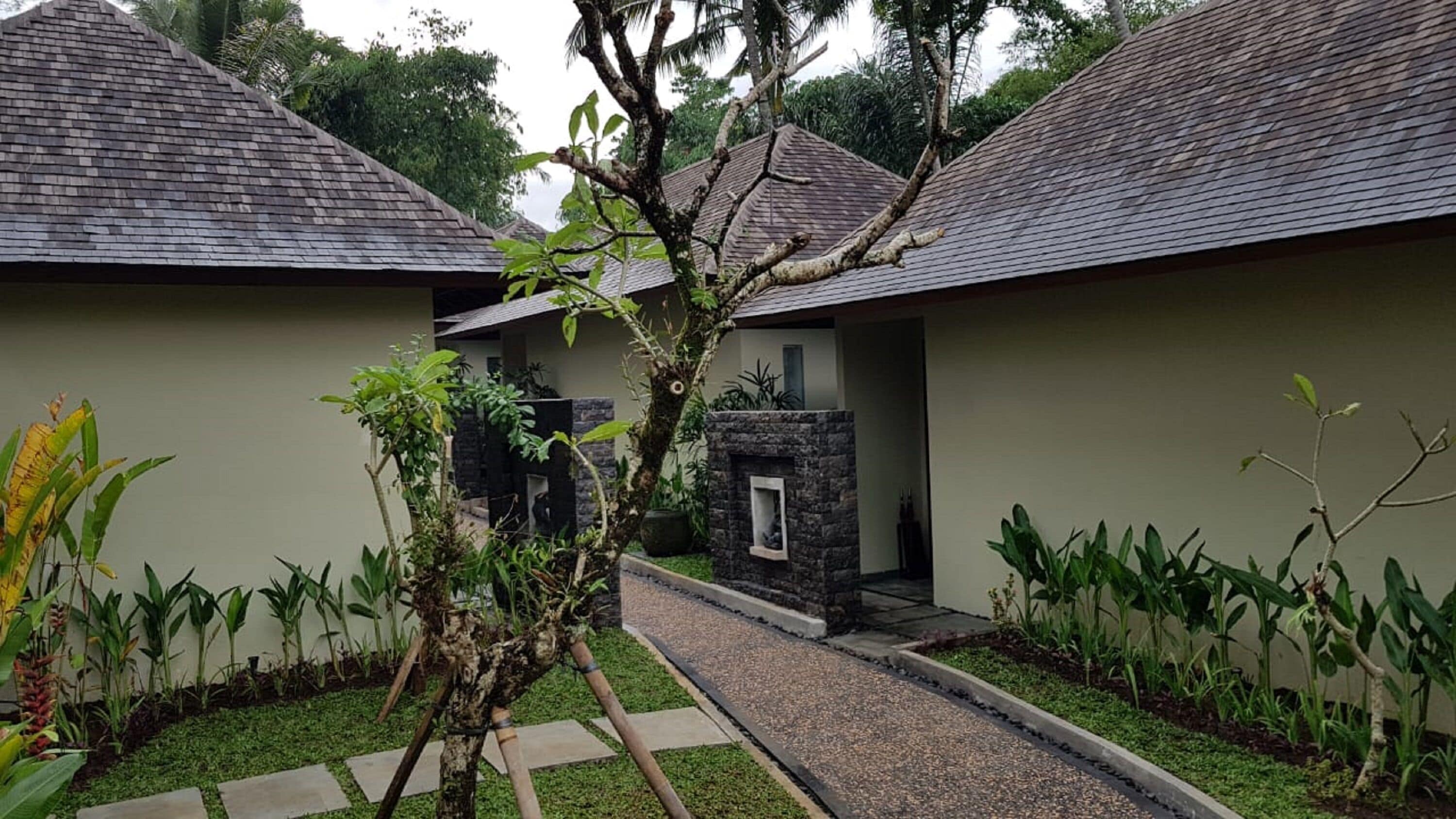 A pathway leading to a building with a stone fireplace, surrounded by lush greenery and a stone wall.