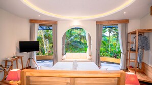 A hotel room with a large bed, a TV, a wooden side table, and a wardrobe. There are two windows with curtains, and a view of greenery outside. - Puri Sebali Resort (Ubud)