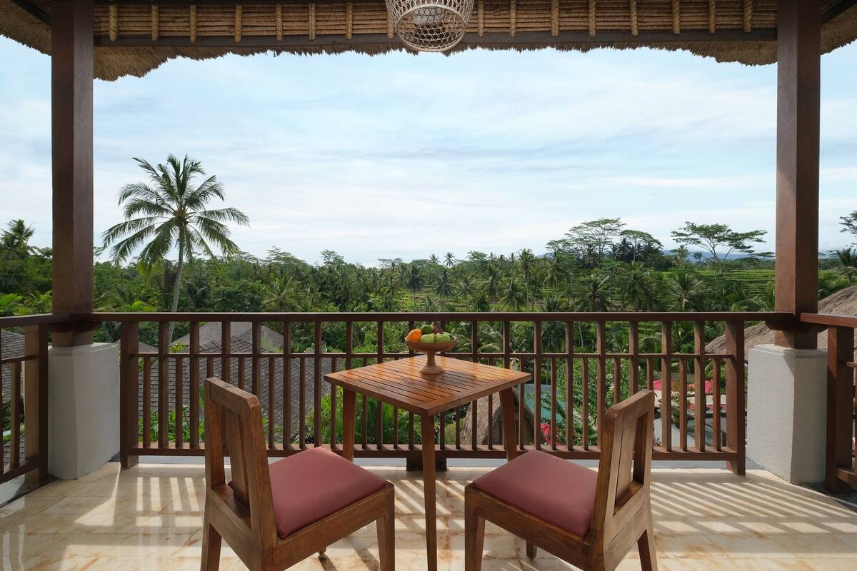 A wooden table with two chairs on a balcony, overlooking a lush landscape with palm trees and a clear sky.