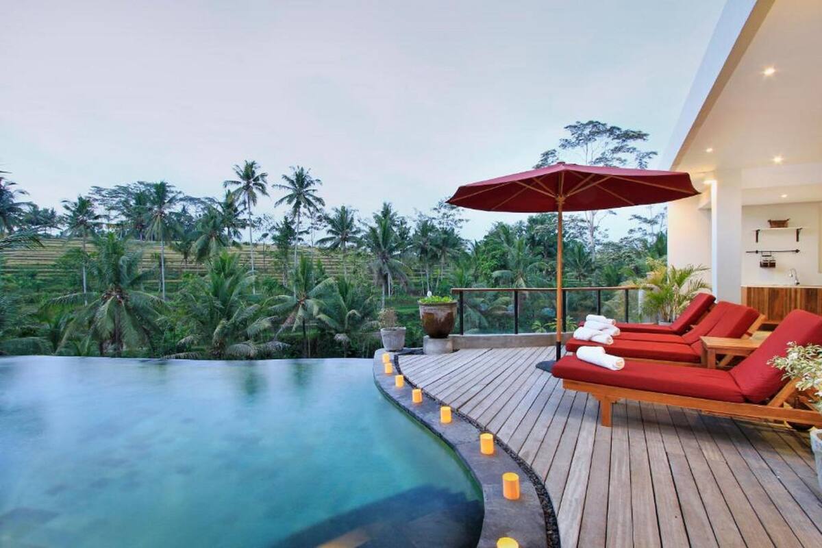 A poolside area with lounge chairs and a red umbrella, surrounded by lush greenery.