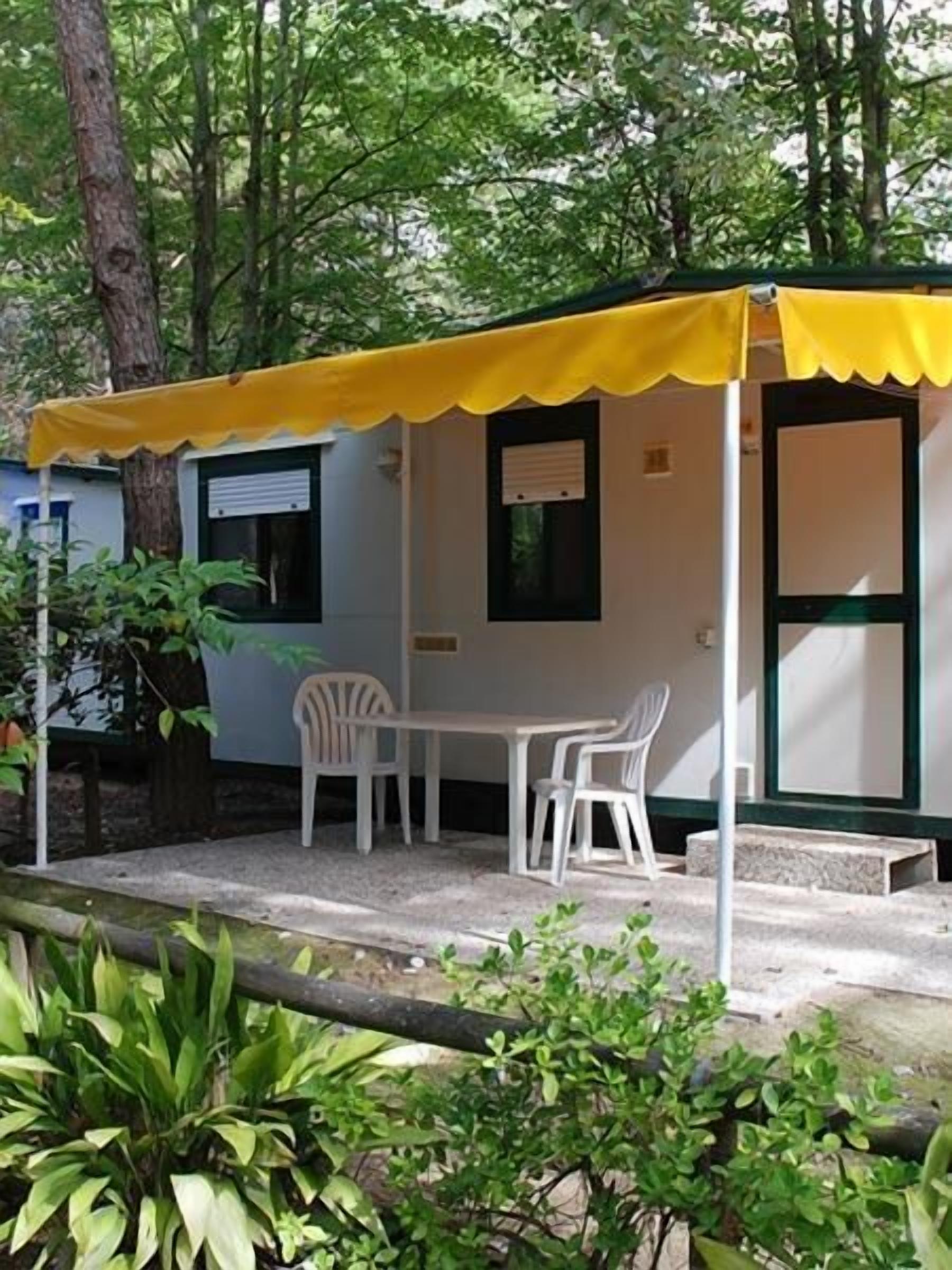 A small, single-story structure with a yellow awning, white walls, and a window with a shutter. There is a white table and two white chairs outside.