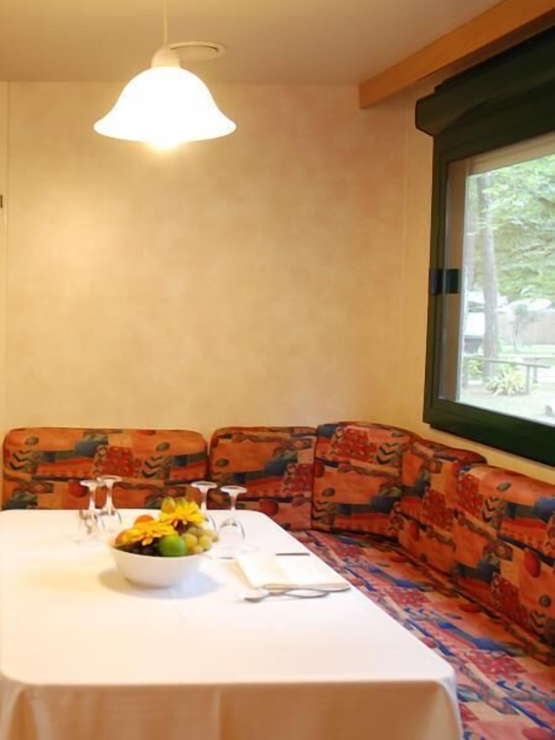 A dining area with a table set for two, a bowl of fruit, and a window with a view of trees.