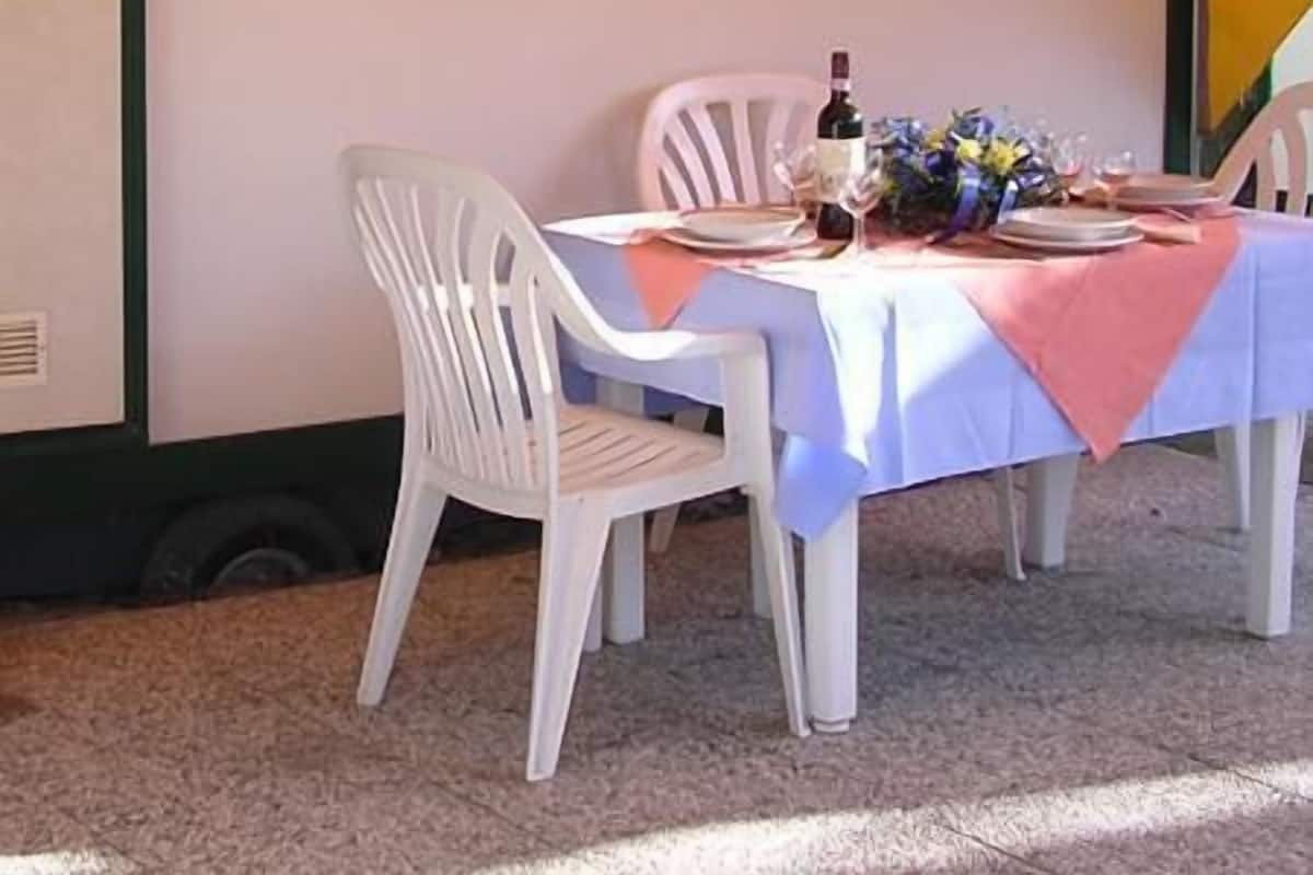 Un espacio al aire libre con comedor, una mesa lista para dos comensales, sillas blancas de plástico y una ventana con cortinas.
