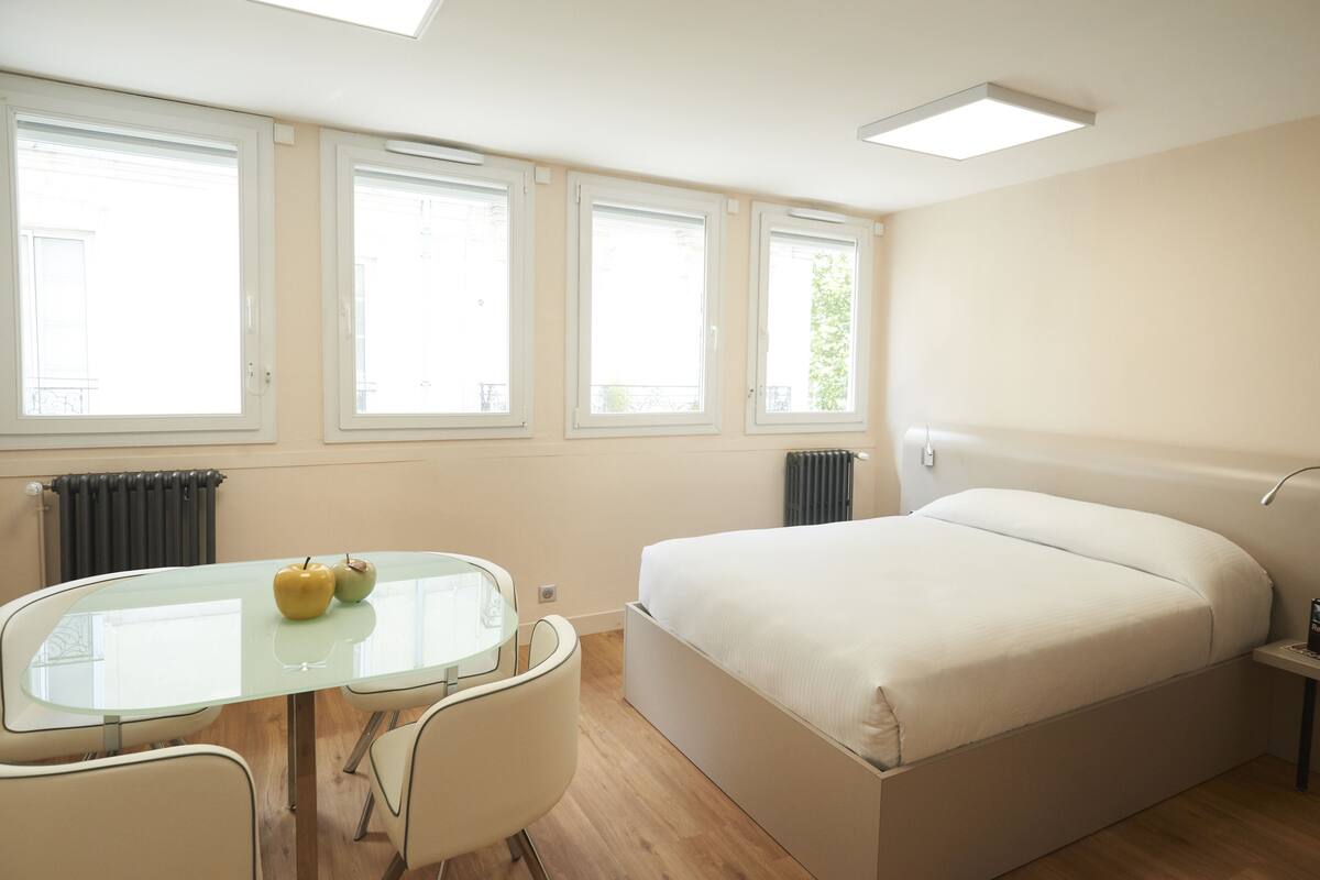 Une chambre d’hôtel moderne avec un grand lit, une table en verre sur laquelle reposent deux pommes, et trois fenêtres laissant entrer la lumière naturelle.