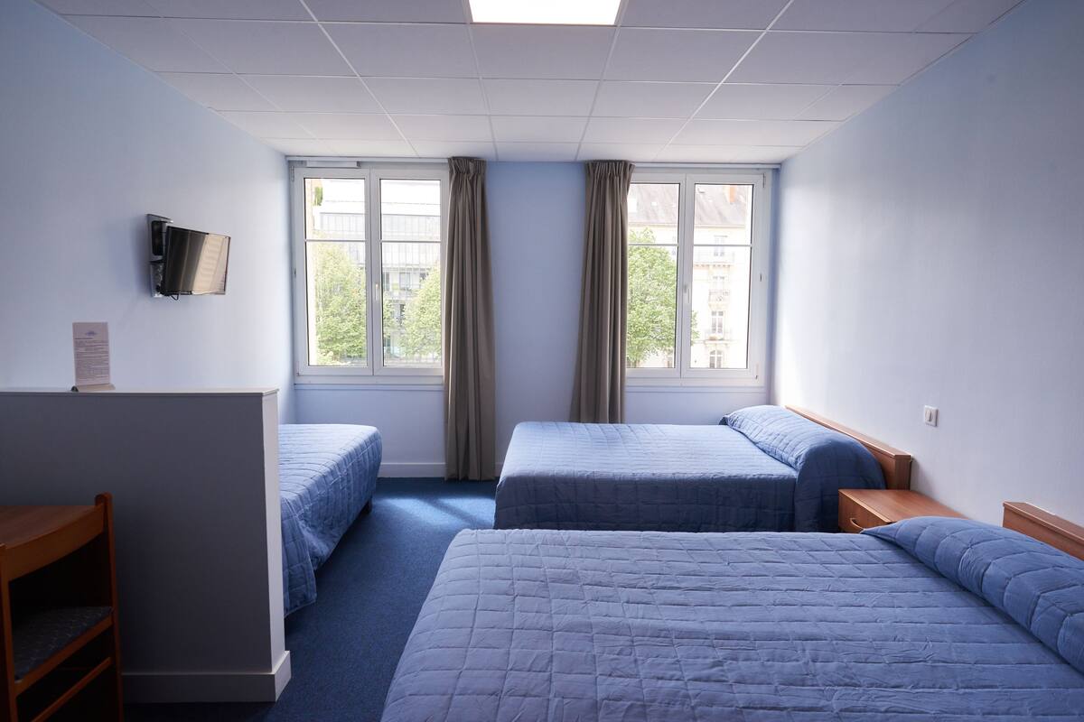 Une chambre d’hôtel avec deux lits simples, un bureau et une chaise. Elle est dotée de fenêtres avec des rideaux, d’un téléviseur à écran plat fixé au mur et d’une petite table en bois.
