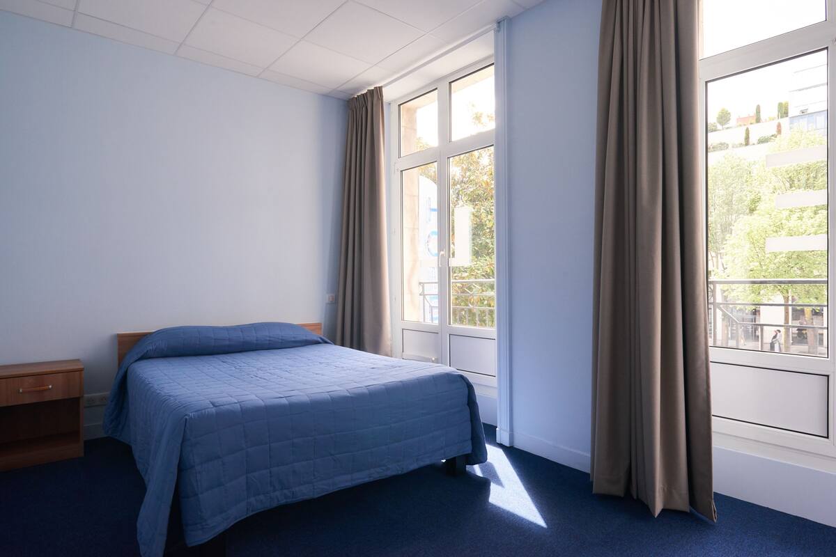 Une chambre d’hôtel avec un lit, une table de chevet, une fenêtre avec des rideaux et une vue sur les bâtiments environnants.