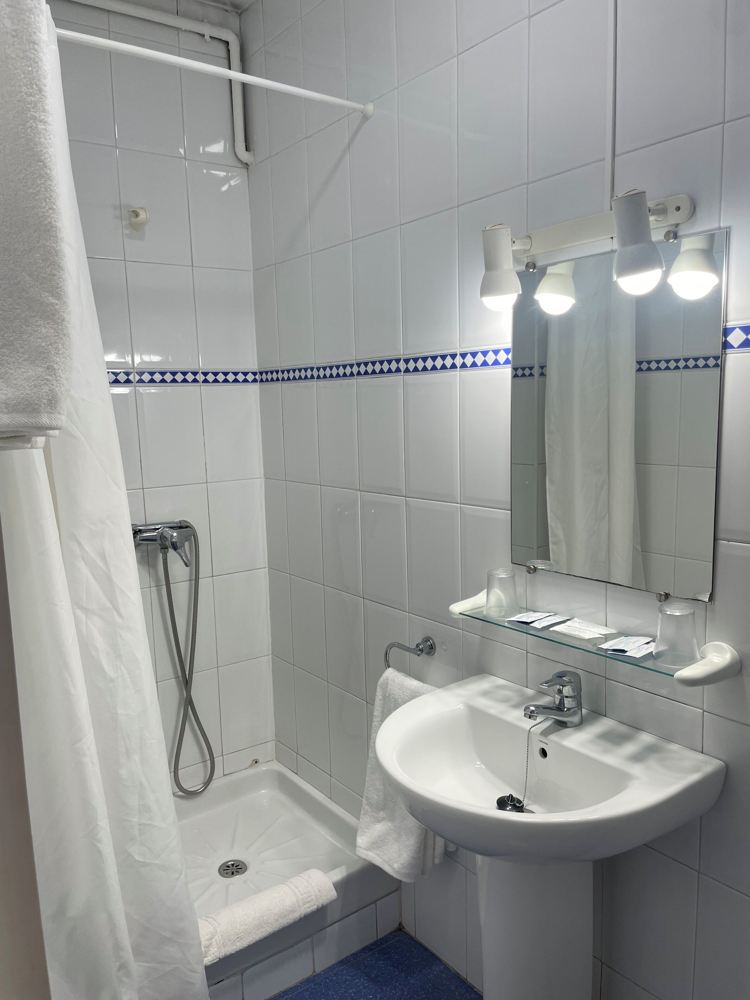 A bathroom with white tiles, a glass shelf with toiletries, a sink, a shower, and a mirror with lights.