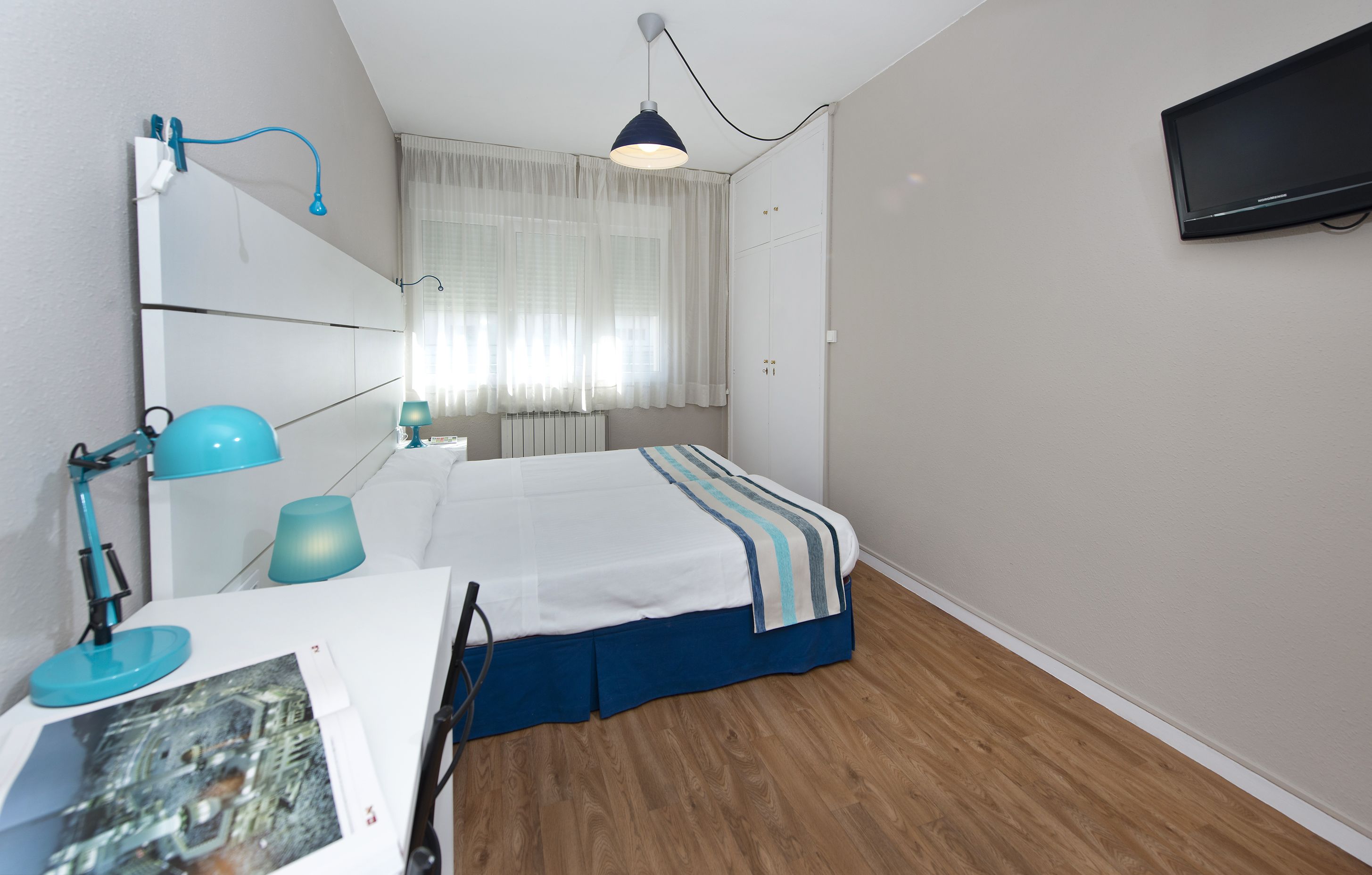 Ein Schlafzimmer mit Bett, Schreibtisch mit Lampe, Wandfernseher und Fenster mit Vorhängen.