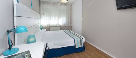 Ein Schlafzimmer mit Bett, Schreibtisch mit Lampe, Wandfernseher und Fenster mit Vorhängen.