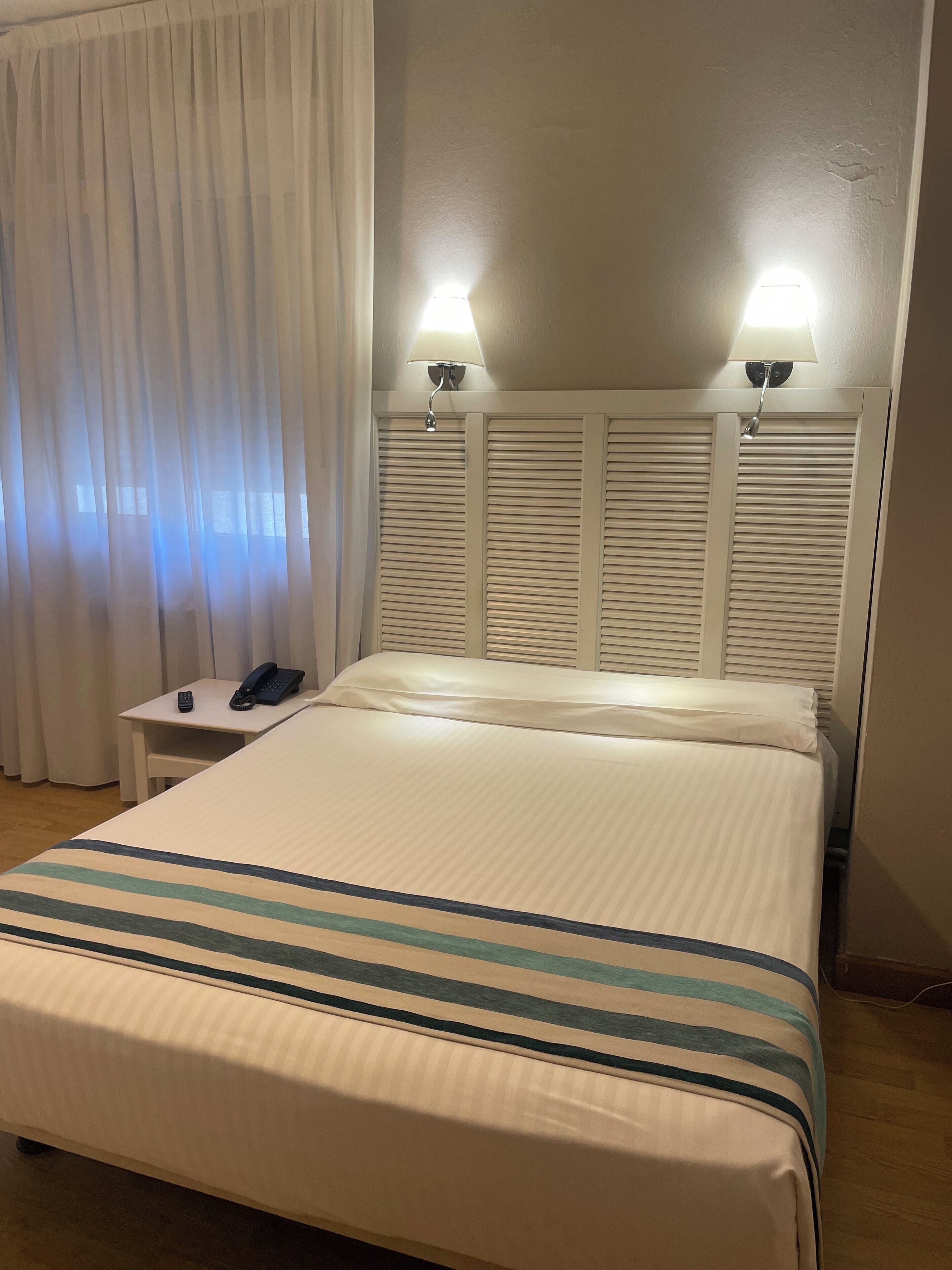 Una habitación de hotel con una cama, dos lámparas de noche, un teléfono junto a la cama y un ventanal con cortinas.
