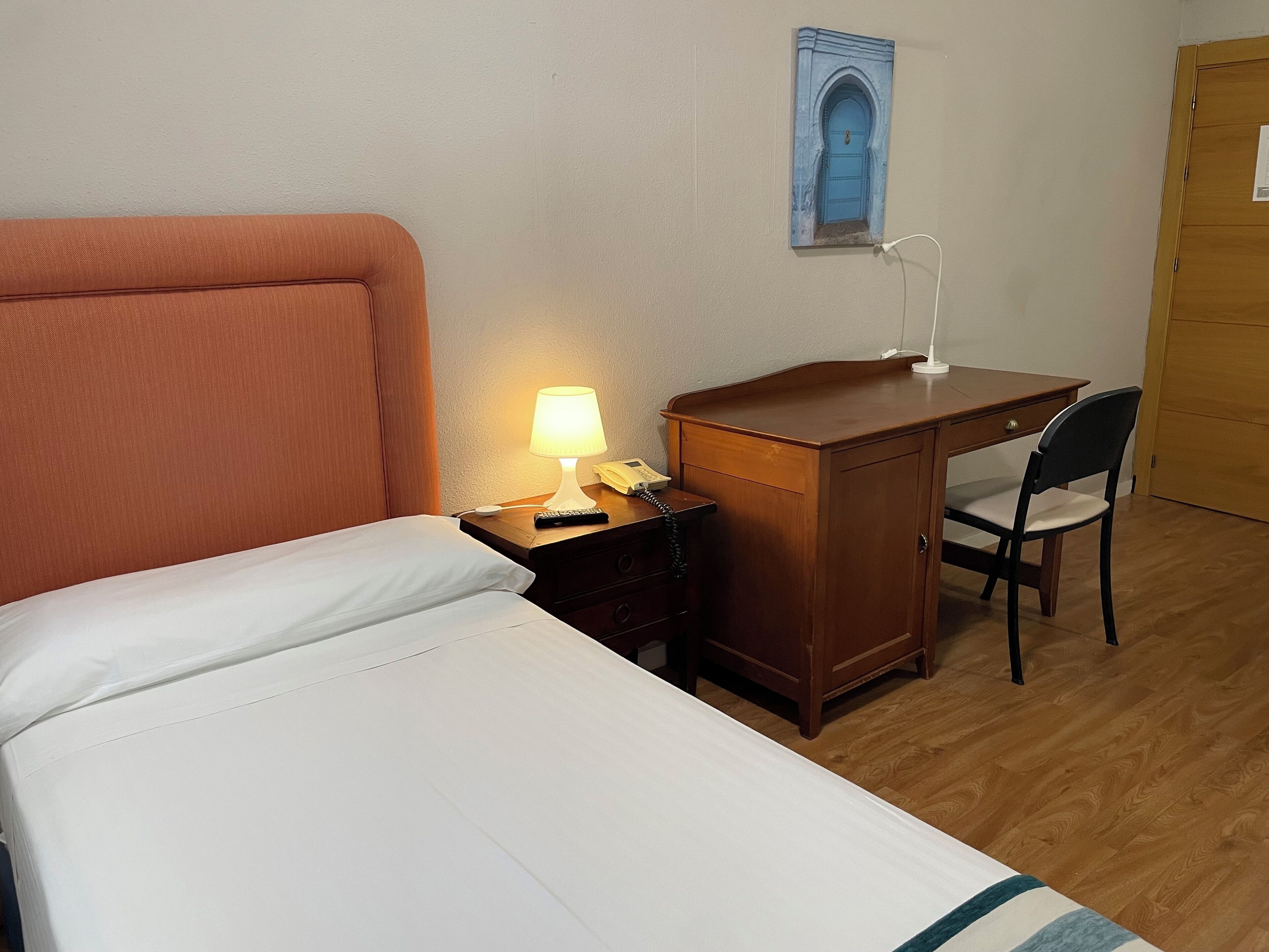 Una habitación de hotel con cama, escritorio, silla, lámpara y una obra de arte en la pared.
