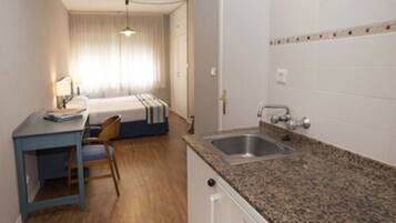 Una habitación de hotel pequeña con cama, escritorio, silla, lavamanos y ventana con cortinas.