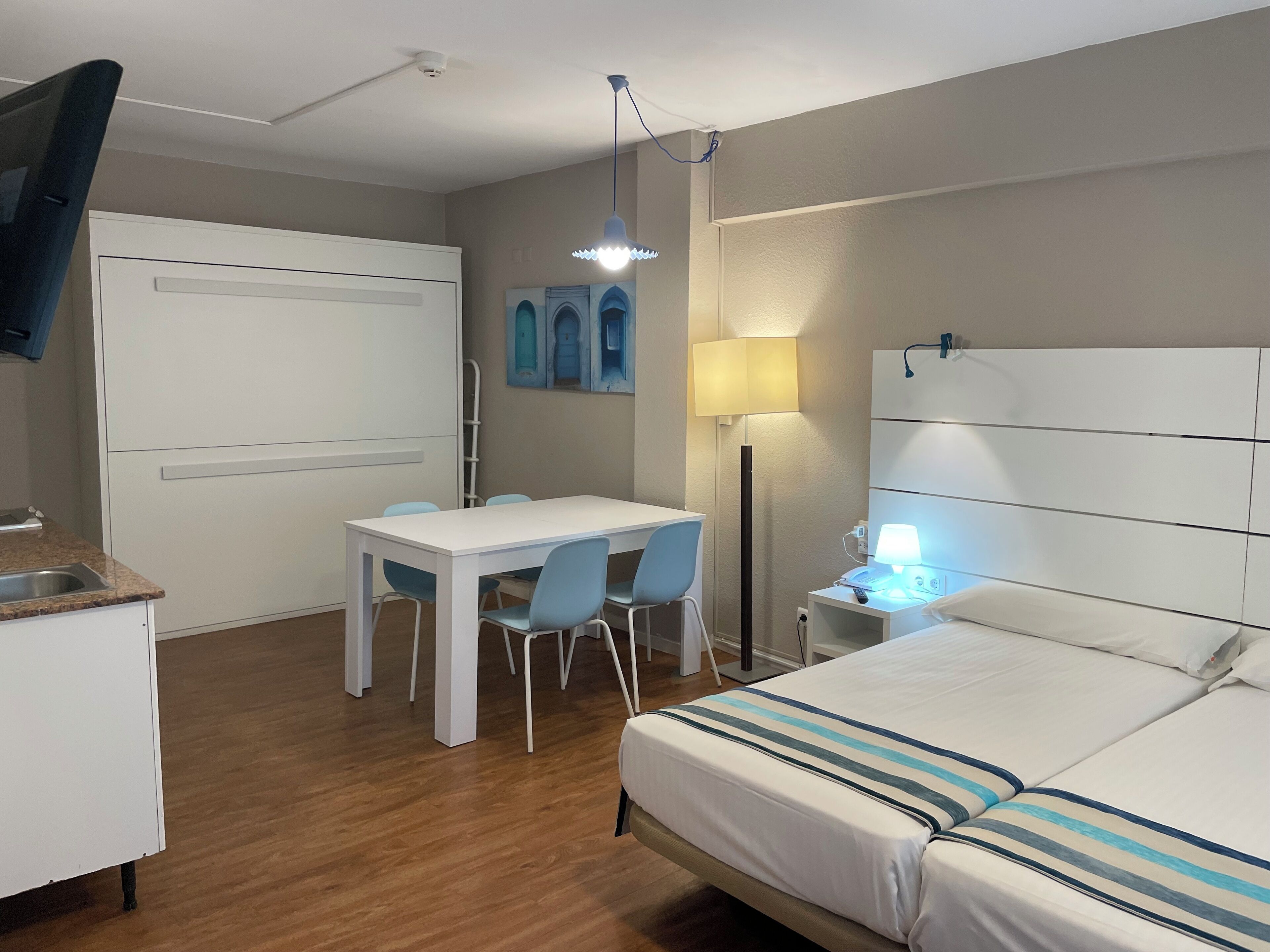 Habitación de hotel con cama, escritorio con sillas, lámpara y estante fijado a la pared.